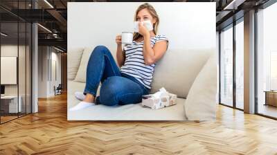 Sick young woman blowing running nose, feeling unhealthy and ill, upset female holding paper napkin, handkerchief, holding tea or coffee mug, sitting on couch at home. Wall mural