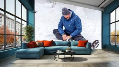 Unhappy young man with injured painful leg sitting on snowy road, outdoors. Portrait of Caucasian young male feeling pain in leg after walking outside in snowy winter park. Young man with ankle injury Wall mural