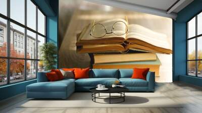 Open book and reading glasses on the table, illuminated by sunlight. Stack of vintage books in the background. Selective focus. Wall mural