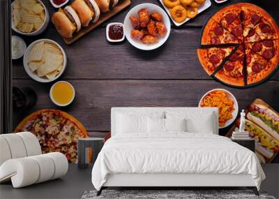 Junk food frame. Pizza, hamburgers, chicken wings, hot dogs and salty snacks. Top view over a dark wood banner background with copy space. Wall mural