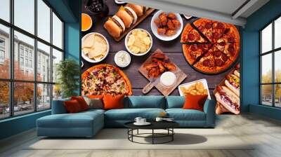 Junk food table scene. Pizza, hamburgers, hot dogs, chicken wings and salty snacks. Overhead view over a dark wood background. Wall mural