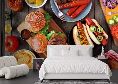 Summer BBQ food table scene with hot dog and hamburger buffet. Top view over a dark wood background. Wall mural