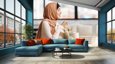 beautiful   smiling muslim woman in traditional religious hijab working remotely behind laptop, communicates by video call . Wall mural