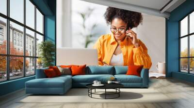 Beautiful young smiling ethnic woman making call via smartphone while working remotely from home while sitting at desk and talking to coworkers . Wall mural