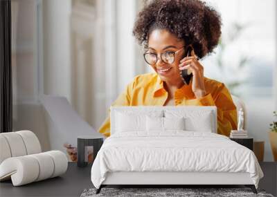 Beautiful young smiling ethnic woman making call via smartphone while working remotely from home while sitting at desk and talking to coworkers Wall mural