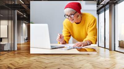 cheerful elderly woman creative designer in red hat smiles at workplace at  table with a laptop, sketches and samples Wall mural