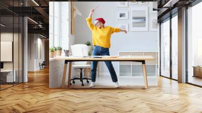 cheerful elderly woman freelancer creative designer in a red hat having fun and dancing in workplace. Wall mural