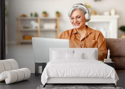 Cheerful elderly woman in headphones using laptop on couch Wall mural