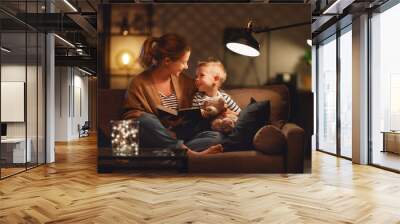 Family before going to bed mother reads to her child son book near a lamp in the evening. Wall mural