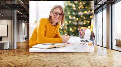 Happy  young woman freelancer laughs while sitting at a workplace with a phone in her hands in a home office working remote  before Christmas. Wall mural