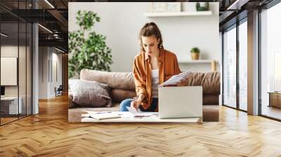 Serious young woman examining papers while working at laptop at home. Wall mural