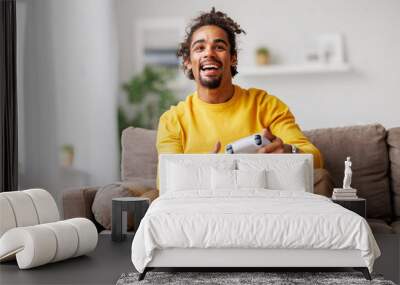 Young positive happy african american guy with wireless gamepad playing video game at home Wall mural