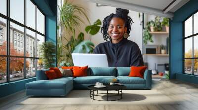 A smiling African American woman working remotely on a laptop with no logo at a desk in a well-lit, organized office space, with a few plants and shelves in the background Wall mural