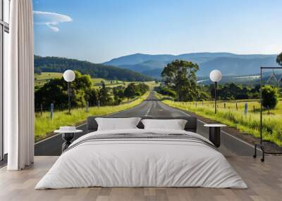 A wide, empty road leading through a scenic landscape with lush green trees and distant mountains under a clear blue sky  Wall mural