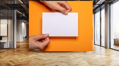 two female hands holding a blank piece of paper with copy space in front of a orange background cardboard Wall mural