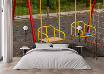 Two yellow swings with red supports stand still on a playground. The ground is made of gravel. There is a net fence behind the swings and green trees in the background Wall mural