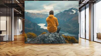 A person is seen from behind, sitting on a rock and gazing at a breathtaking landscape. The individual is wearing a mustard yellow jacket with a hood, dark pants, and sneakers. The jacket’s hood is up Wall mural