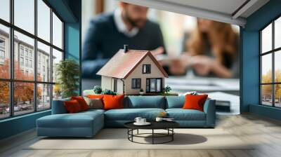 Close-up of a miniature house model and keys, with a real estate agent and a couple signing a contract in the blurred background. Wall mural