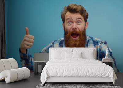 A bearded man working in a cozy café, wearing casual attire and using a laptop, pointing at the copy space with an astonished and happy expression, against a soft, sky blue background Wall mural