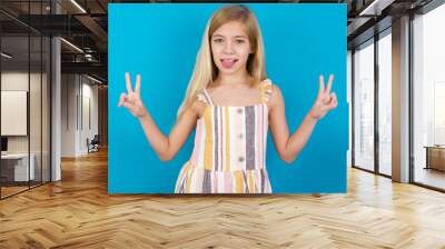 beautiful Caucasian little girl wearing stripped dress over blue background with optimistic smile, showing peace or victory gesture with both hands, looking friendly. V sign. Wall mural