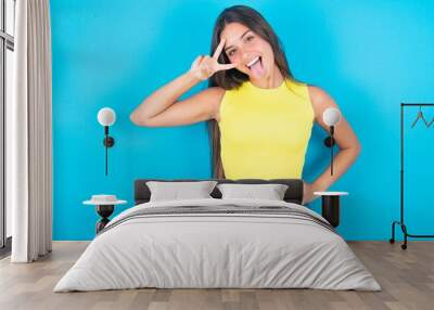 brunette woman wearing yellow tank top over blue background making v-sign near eyes. Leisure, coquettish, celebration, and flirt concept. Wall mural