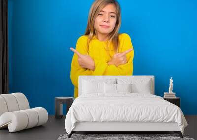 caucasian teen girl wearing yellow sweater over blue studio background crosses arms and points at different sides hesitates between two items or variants. Needs help with decision Wall mural