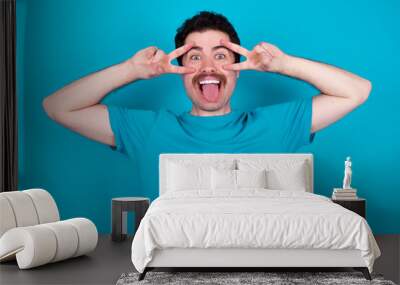 Cheerful positive young handsome Caucasian man with moustache wearing blue t-shirt against blue background shows v-sign near eyes open mouth Wall mural