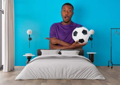 Confused Man wearing purple T-shirt holding a ball over blue background chooses between two ways, points at both sides with crossed hands, feels doubt. Need your advice. Wall mural