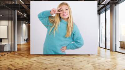 Cute Caucasian kid girl wearing blue knitted sweater against white wall making v-sign near eyes. Leisure, coquettish, celebration, and flirt concept. Wall mural