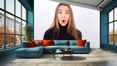 Emotional attractive Young caucasian girl wearing black turtleneck over white wall with opened mouth expresses great surprise and fright, stares at camera. Unexpected shocking news and human reaction. Wall mural