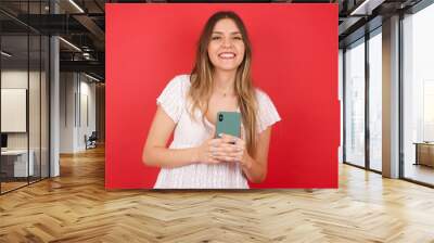 Girl turn attention at camera, distracted from smartphone smiling friendly and happily holding mobile phone as, taking selfie in mirror, showing friends new outfit, standing in dressing room of mall. Wall mural