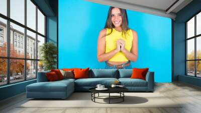 Happy brunette woman wearing yellow tank top over blue background stands against orange studio wall keeps hands on heart, swears be loyal, expresses gratitude. Honesty concept. Wall mural