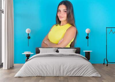 Image of cheerful brunette woman wearing yellow tank top over blue background with arms crossed. Looking and smiling at the camera. Confidence concept. Wall mural