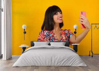 Isolated shot of pleased cheerful young woman with dark hair, makes selfie with mobile phone, dressed in casual sweater, isolated over beige background. People, technology and leisure concept Wall mural