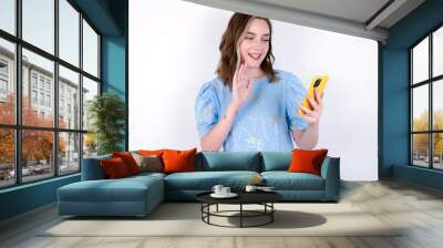 Portrait of happy friendly young caucasian woman wearing blue T-shirt over white background taking selfie and waving hand, communicating on video call, online chatting. Wall mural