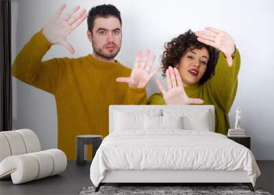 Portrait of smiling Young couple wearing knitted sweater standing against white wall looking at camera and gesturing finger frame. Creativity and photography concept. Wall mural