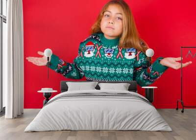 Puzzled and clueless little kid girl wearing knitted sweater christmas over red background with arms out, shrugging shoulders, saying: who cares, so what, I don't know. Negative human emotions. Wall mural