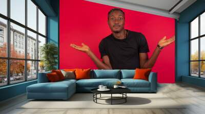 Puzzled and clueless Young African American handsome man standing against red background with arms out, shrugging shoulders, saying: who cares, so what, I don't know. Negative human emotions. Wall mural