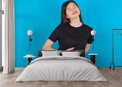 Satisfied smiling young caucasian woman wearing black T-shirt over blue, keeps hands on belly, being in good mood after eating delicious supper, demonstrates she is full. Pleasant feeling in stomach. Wall mural