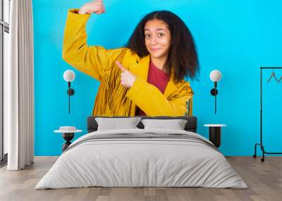 Smiling African teenager girl wearing yellow jacket over blue background raises hand to show muscles, feels confident in victory, strong and independent. Wall mural