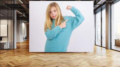 Smiling Cute Caucasian kid girl wearing blue knitted sweater against white wall raises hand to show muscles, feels confident in victory, strong and independent. Wall mural