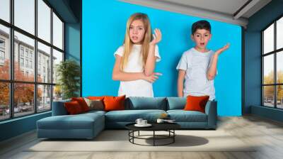 Studio shot of frustrated two kids boy and girl standing over blue studio background gesturing with raised palm, frowning, being displeased and confused with dumb question. Wall mural