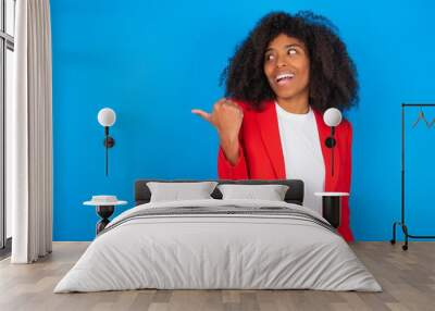 Stupefied young businesswoman with afro hairstyle wearing red over blue with surprised expression, opens eyes and mouth widely, points aside with thumb, shows something strange. Advertisement concept. Wall mural