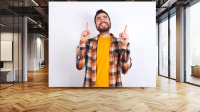 Successful friendly looking young caucasian man wearing plaid shirt over white background exclaiming excitedly, pointing both index fingers up, indicating something. Wall mural