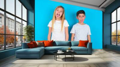 two kids boy and girl standing over blue studio background with happy and funny face smiling and showing tongue. Wall mural