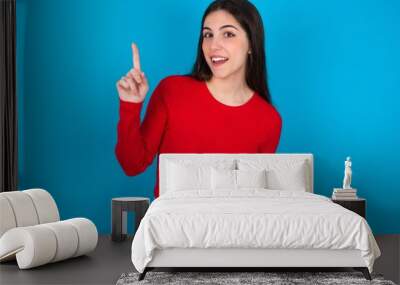 young brunette girl wearing red T-shirt against blue wall pointing finger up and looking inspired by genius thought, showing good idea sign, having clever solution in mind Wall mural