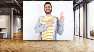 Young caucasian mán wearing trendy clothes over white background smiling swearing with hand on chest and fingers up, making a loyalty promise oath. Wall mural