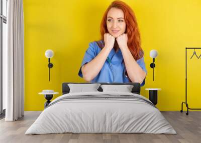 young red-haired doctor woman over yellow studio background holds hands under chin, glad to hear heartwarming words from stranger Wall mural