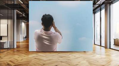 Back view of man using cell phone to take photo with cloud and sky in background. Asian male rests their eyes while looking up at beautiful sky. Man use mobile phone to take selfie. Outdoor happiness. Wall mural