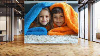 Two children smiling under colorful blankets on a cozy carpet. Wall mural